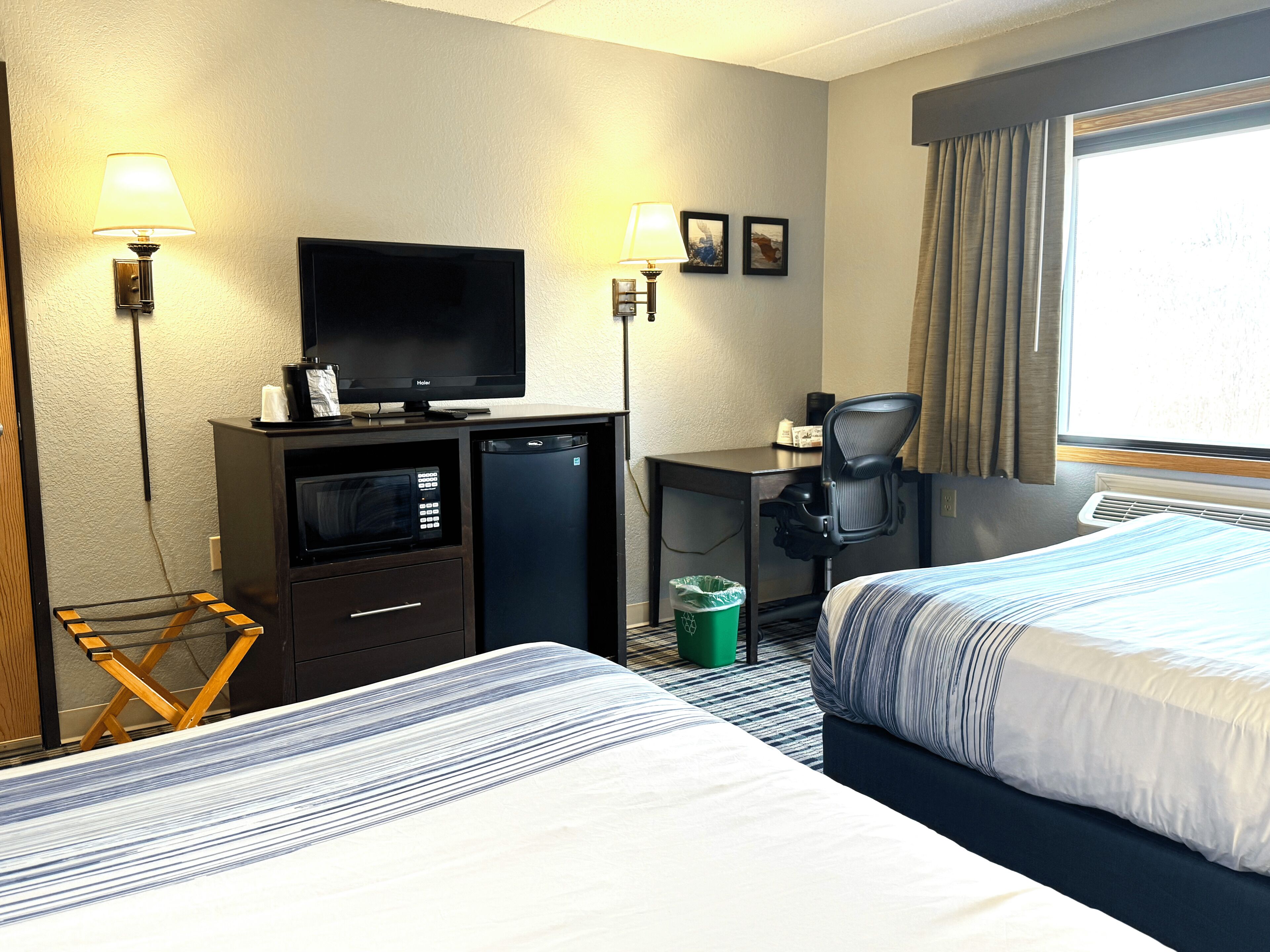 A hotel room with two beds, a desk with a chair, a television, a microwave, and a trash bin.