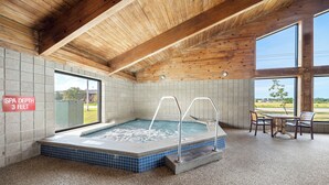 A spa area with a hot tub, wooden ceiling beams, tiled walls, and a view through large windows. - AmericInn by Wyndham Ames (Ames)