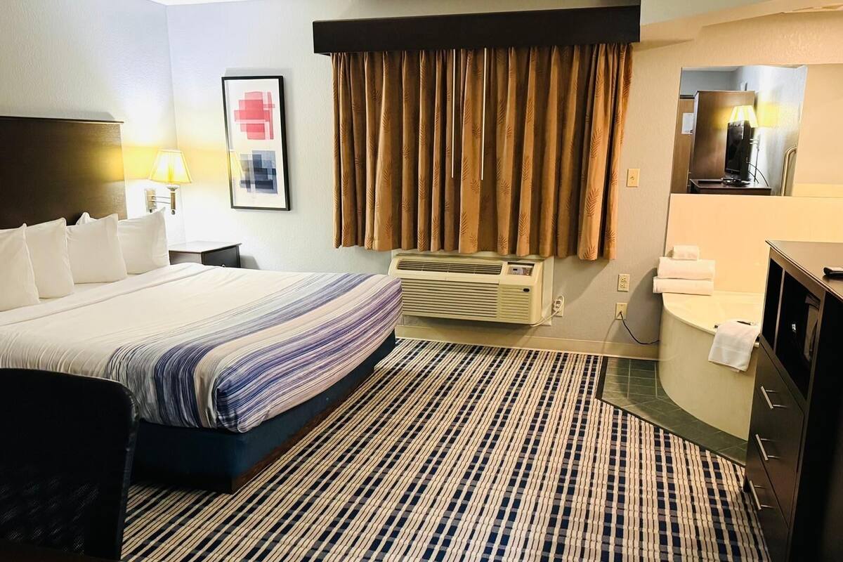 A hotel room with a bed, a desk, a chair, a desk lamp, a wall-mounted TV, a bathroom with a bathtub, and a patterned rug.