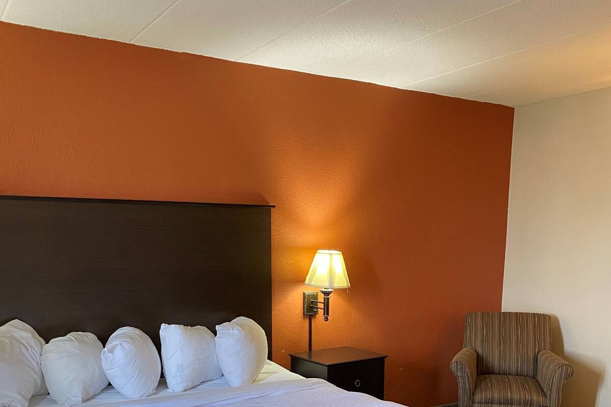 A hotel room with a large bed, a nightstand with a lamp, a striped armchair, and a red ottoman.