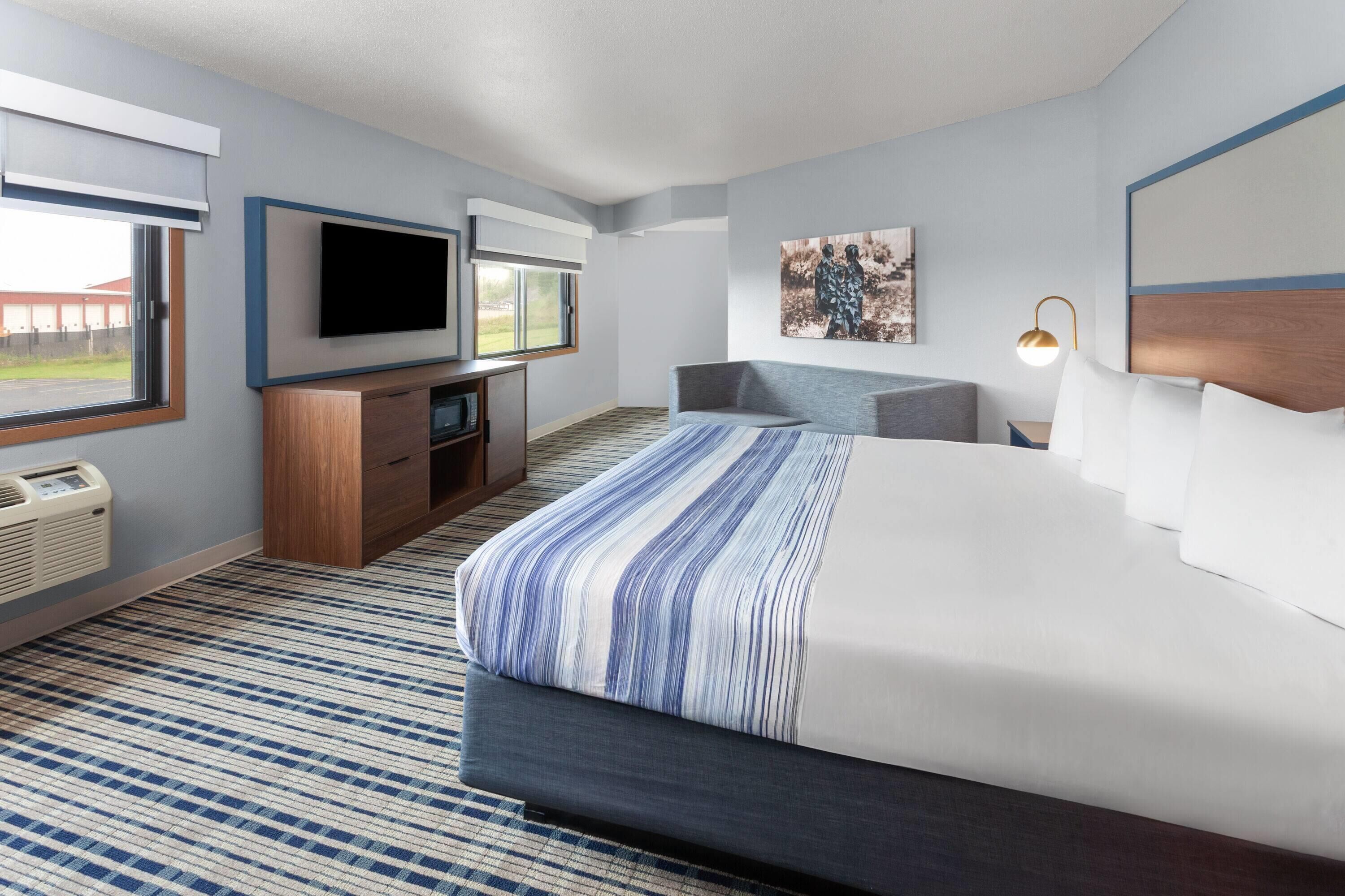 A hotel room with a large bed, a flat-screen TV mounted on the wall, a desk with a chair, and a window with a view of the outside.