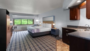 Ein modernes Hotelzimmer mit einem großen Bett, einem Schreibtisch, einem Sessel, einem Fenster mit Blick auf Grünflächen, einer Küchenzeile mit Spüle und Arbeitsplatte sowie einem gemusterten Teppich.