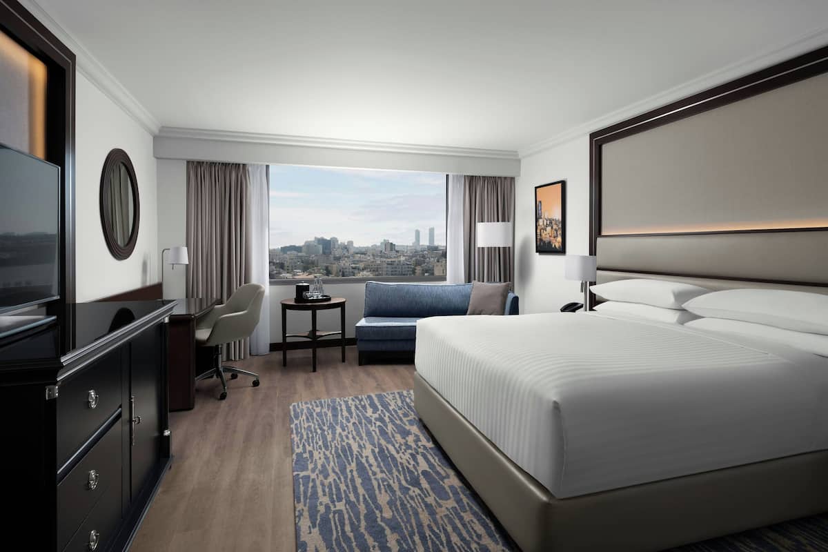 Ein Hotelzimmer mit einem großen Bett, einem Schreibtisch, einem Sessel, einem Fernseher und Blick auf die Stadt.