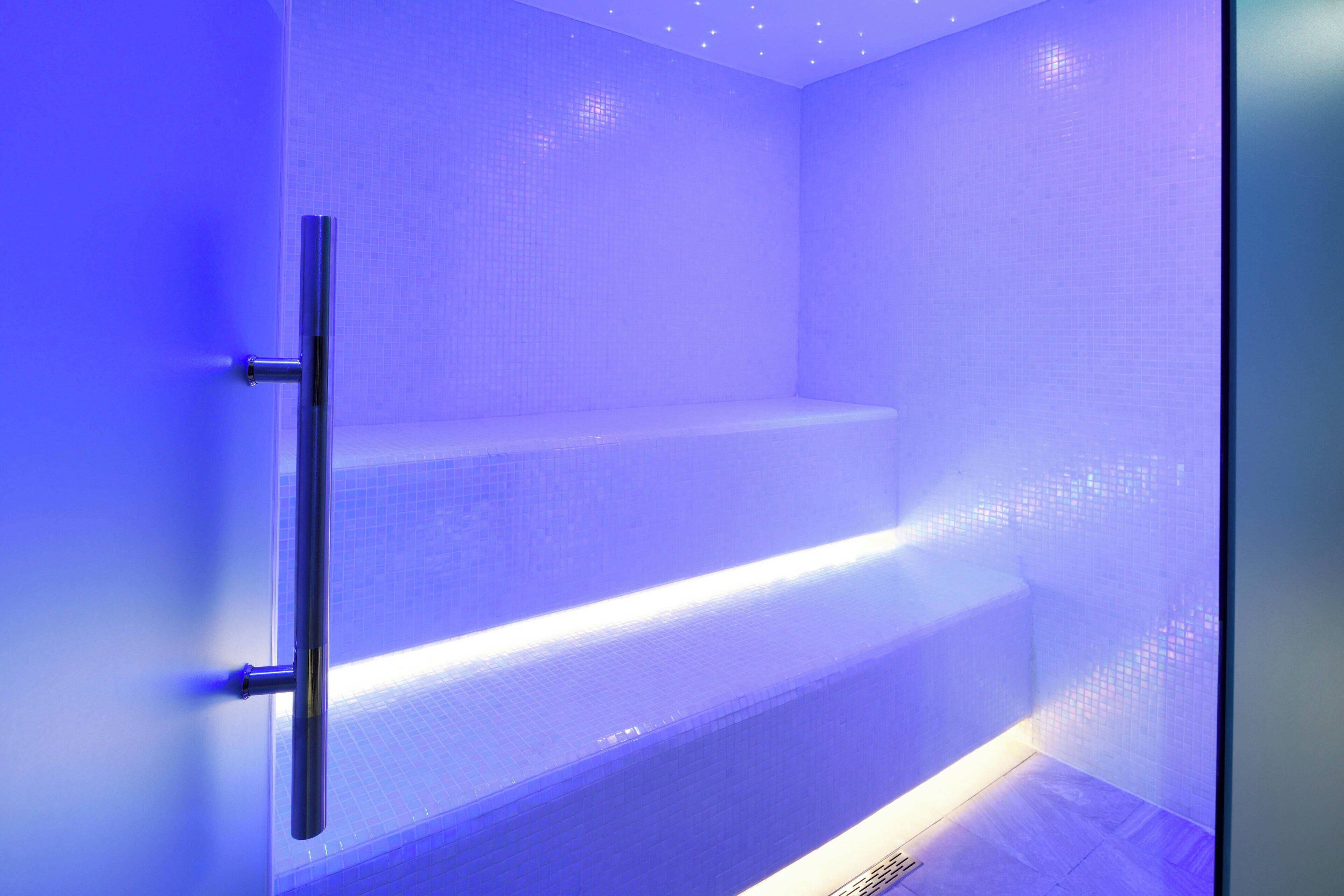 Un baño con ducha iluminada en azul, mampara de vidrio y paredes de azulejos.