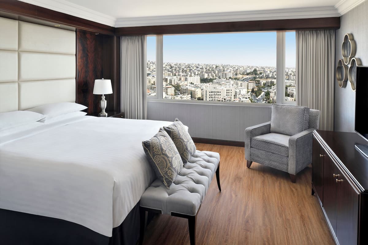 Ein Hotelzimmer mit einem großen Bett, einer Sitzecke, einem Sessel, einem Fernseher und Blick auf eine Stadtlandschaft.