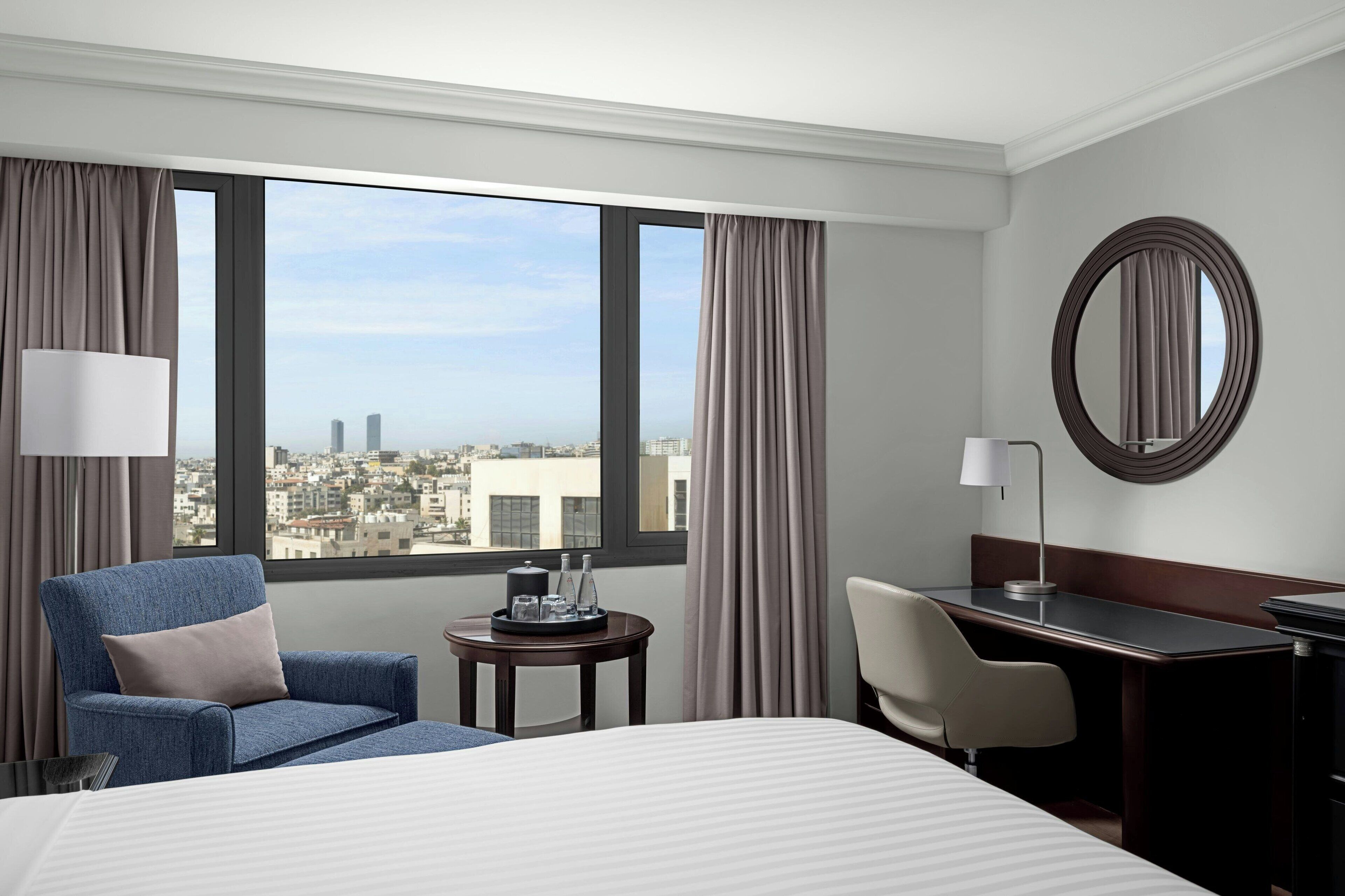 A hotel room with a large bed, a desk, a chair, a round mirror, and a view of the city through large windows.