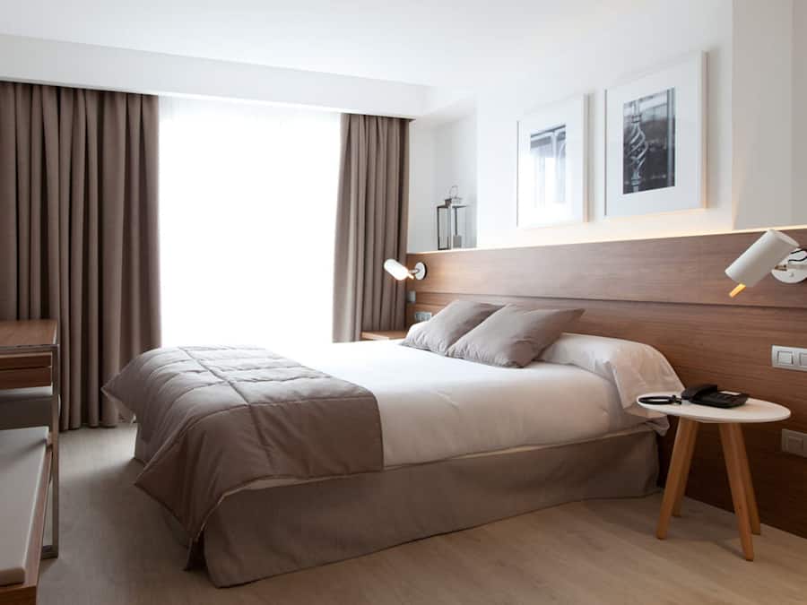 A modern hotel room with a large bed, a desk, a chair, a small table, and a bedside lamp.