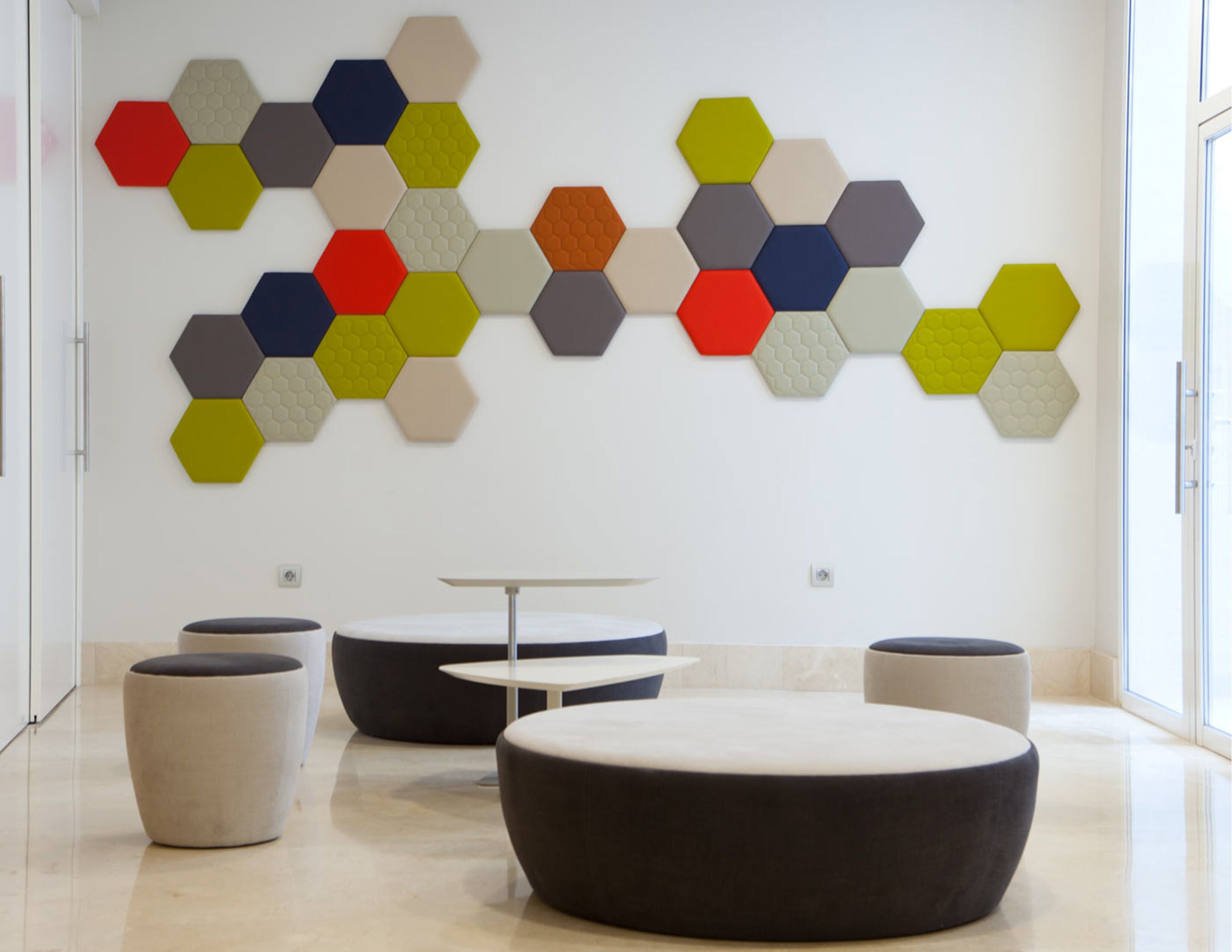 Un interior moderno con arte mural hexagonal, un otomano negro redondo y una pequeña mesa blanca.
