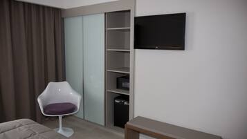A modern hotel room with a bed, a seating area with a chair, a TV mounted on the wall, and a wardrobe with sliding doors.