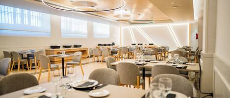A modern dining area with multiple tables and chairs, large windows, and contemporary lighting fixtures.