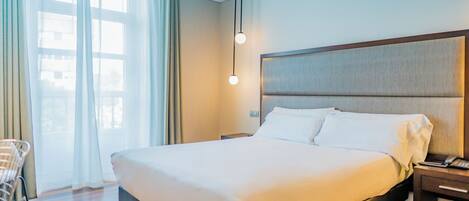 Habitación de hotel con una cama grande, una mesita de noche con lámpara y una ventana con cortinas.