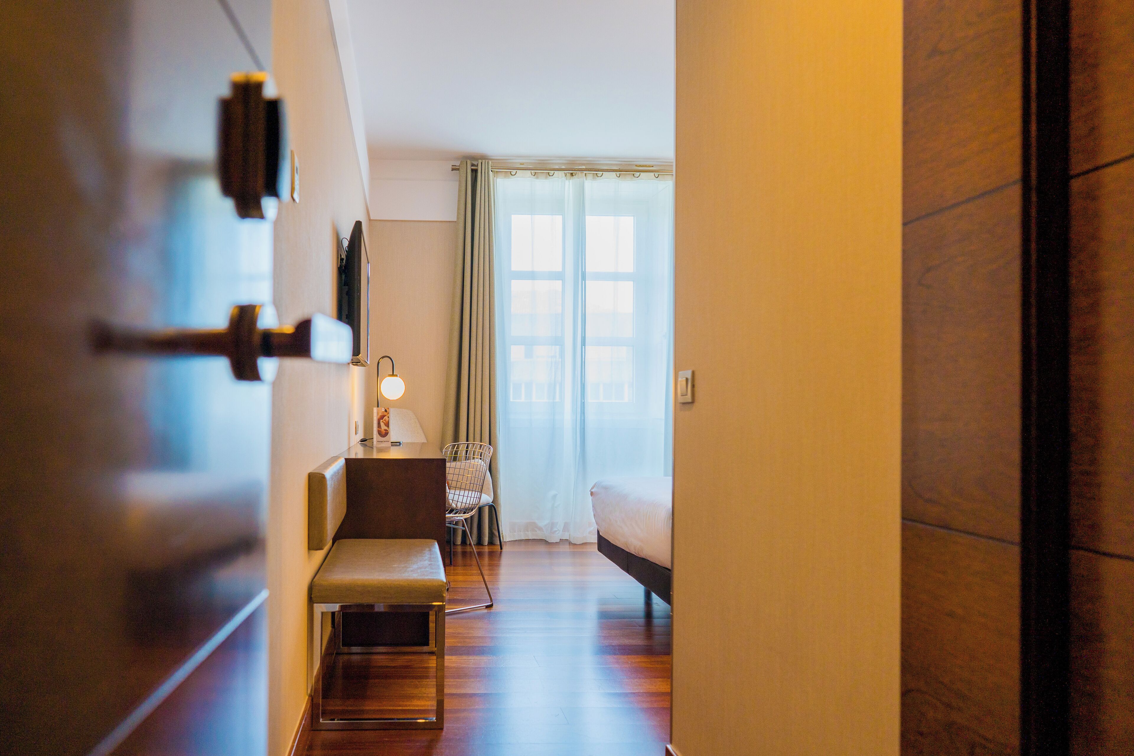 A hotel room with a wooden floor, a bed, a bench, a chair, a desk with a lamp, and a window with curtains.