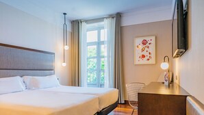 Habitación de hotel con una cama grande, un escritorio, una silla, una ventana con cortinas y un televisor instalado en la pared.