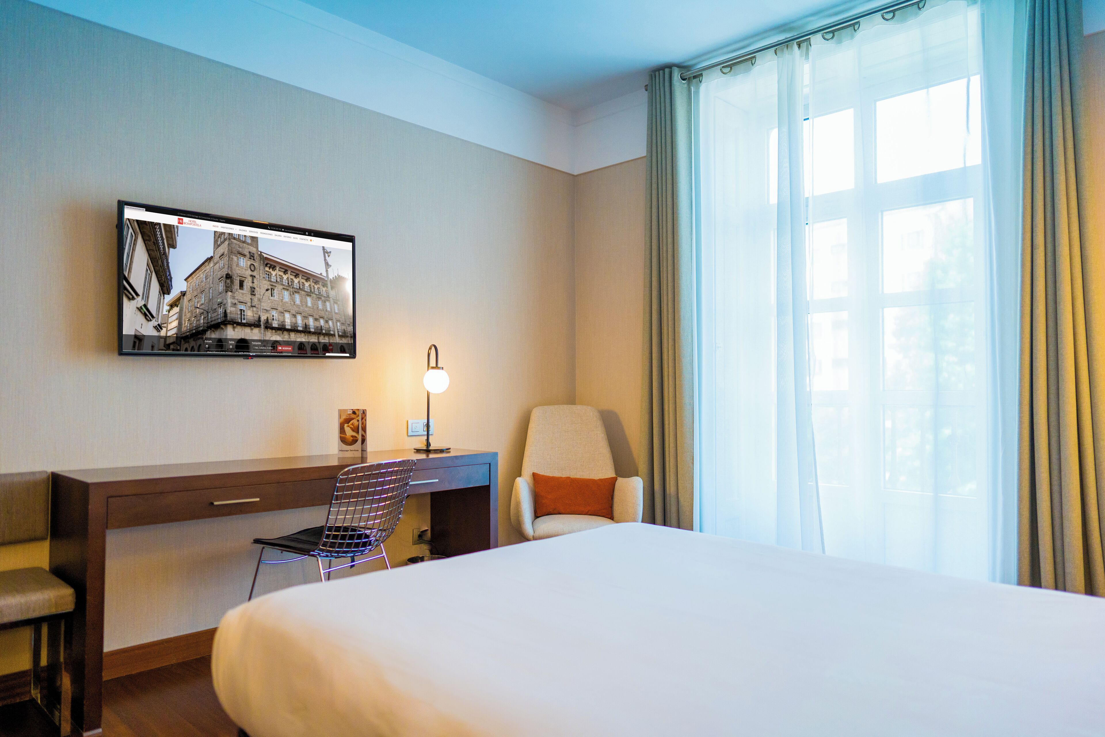 Une chambre d’hôtel comprenant un lit, un bureau, une chaise, un téléviseur fixé au mur et une fenêtre avec des rideaux.
