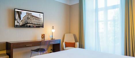 Une chambre d’hôtel comprenant un lit, un bureau, une chaise, un téléviseur fixé au mur et une fenêtre avec des rideaux.