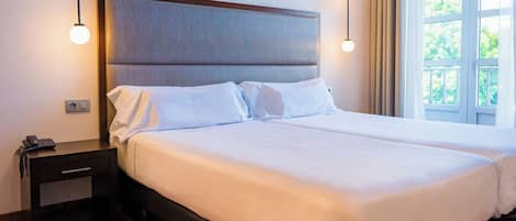 Una cama doble con cabecera de madera, una mesita de noche con un teléfono y una ventana con cortinas.