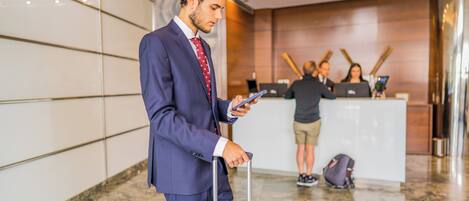 A man in a blue suit holding a phone and a suitcase in a hotel lobby with a reception desk and staff in the background.