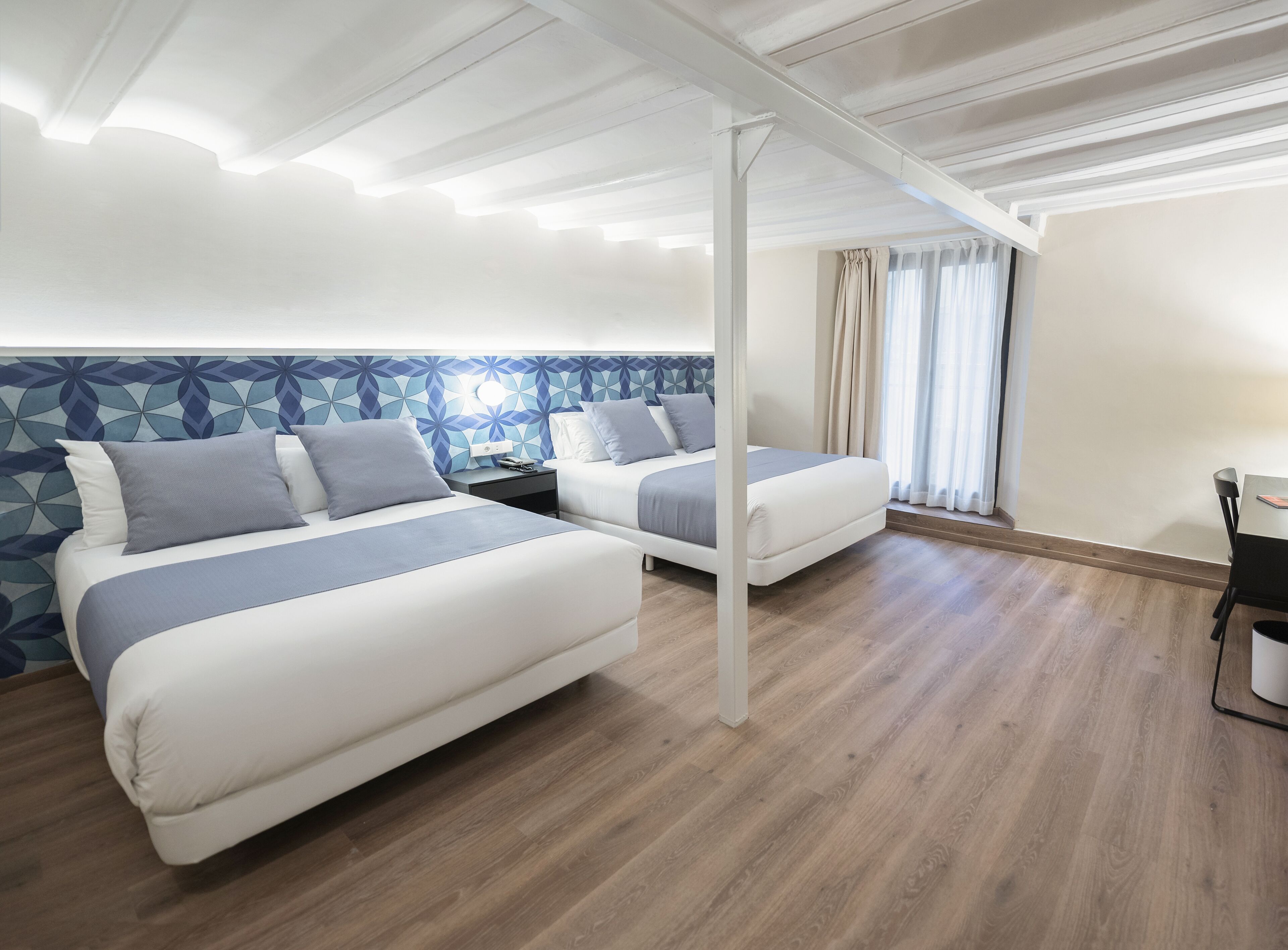 A hotel room with two beds, a wooden floor, a patterned wall, and a window with curtains.