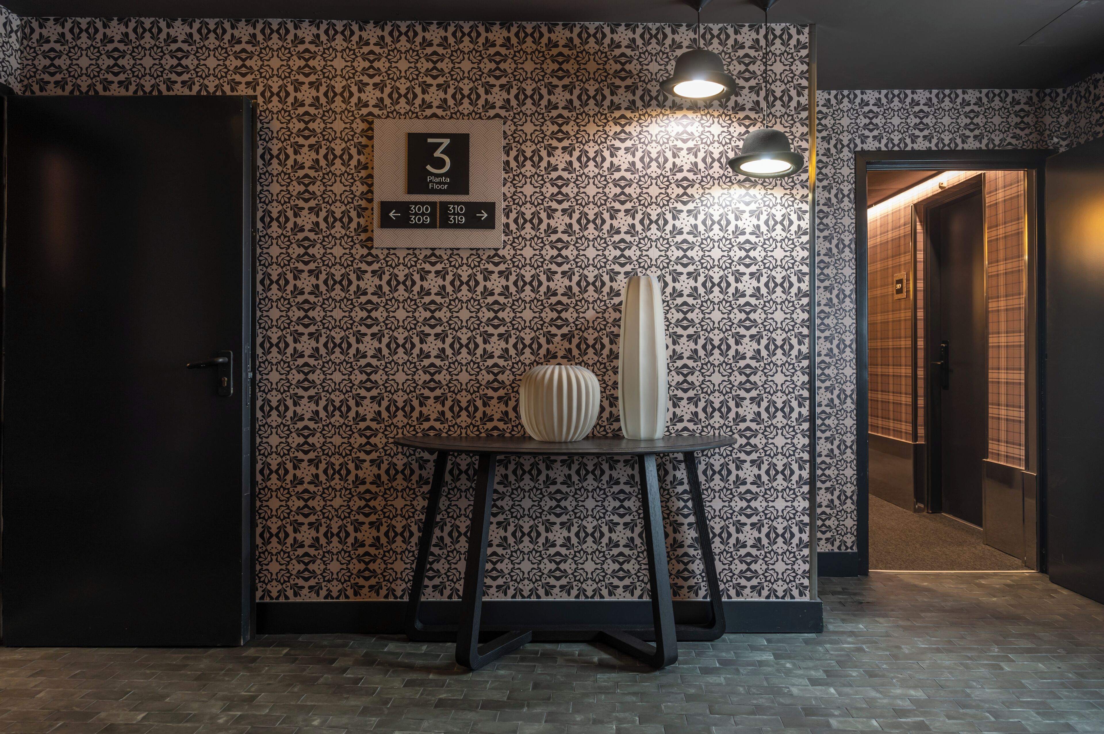 A hotel interior with a patterned wall, a sign indicating the 3rd floor, a table with two vases, and a door to the left.