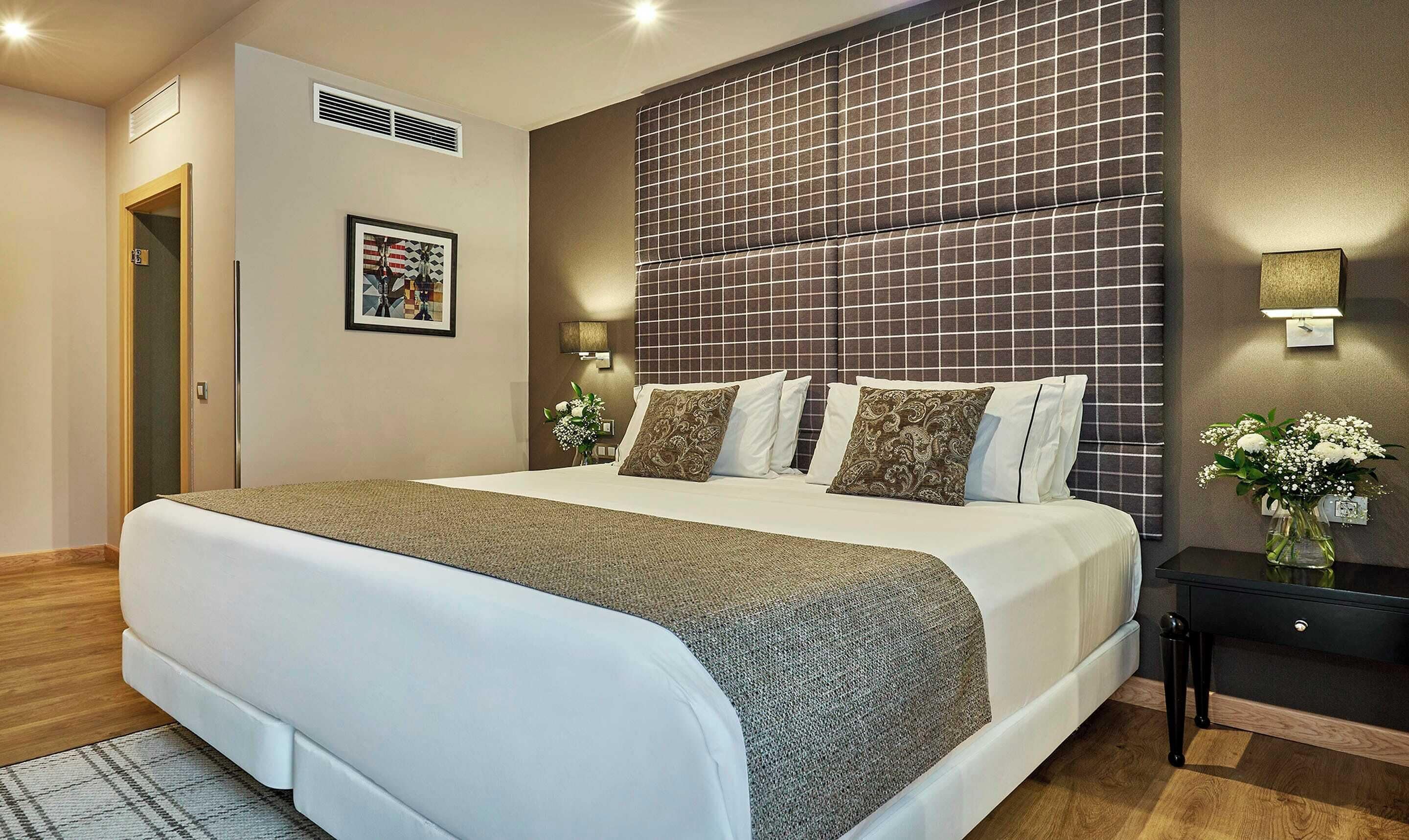 Habitació d'hotel amb un llit gran, una tauleta de nit amb un vas de flors i un llum penjada a la paret.