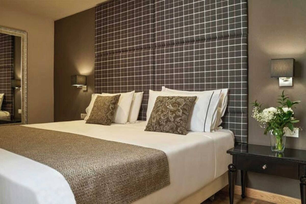 A hotel room with a large bed, a nightstand, a mirror, a wall-mounted light, and a vase with flowers.