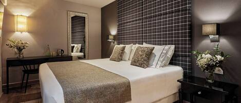 A hotel room with a large bed, a desk with a chair, a small table, and a wall-mounted lamp.