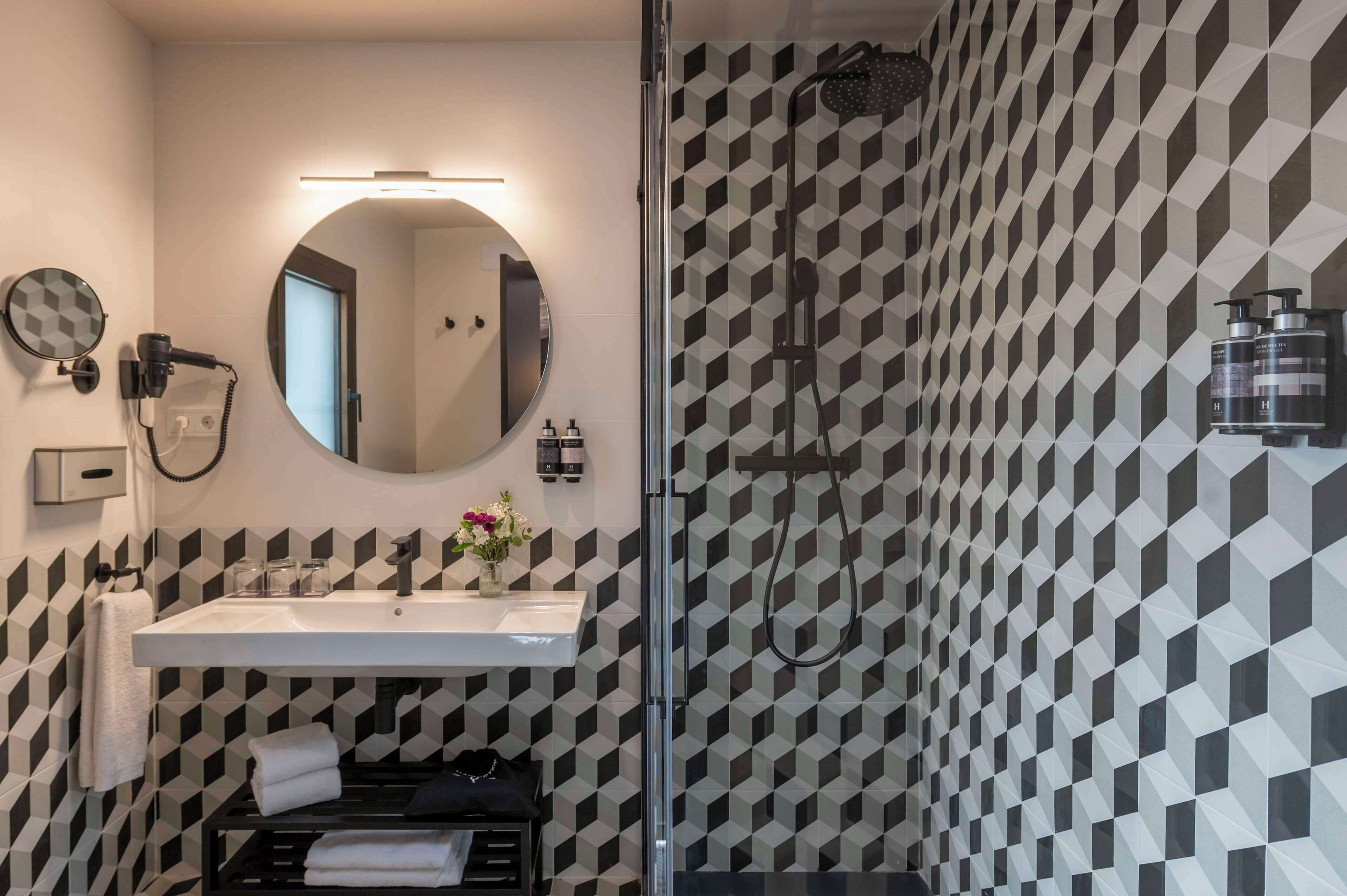 Une salle de bain moderne avec un mur orné d’un motif géométrique, un miroir rond, un lavabo et une douche.