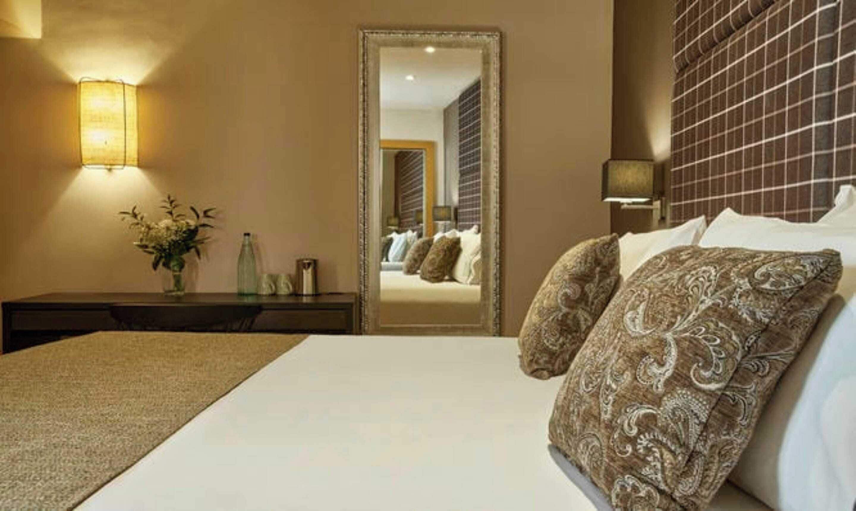 A hotel room with a large bed, a bedside table with a vase of flowers, a mirror, and a wall-mounted lamp.