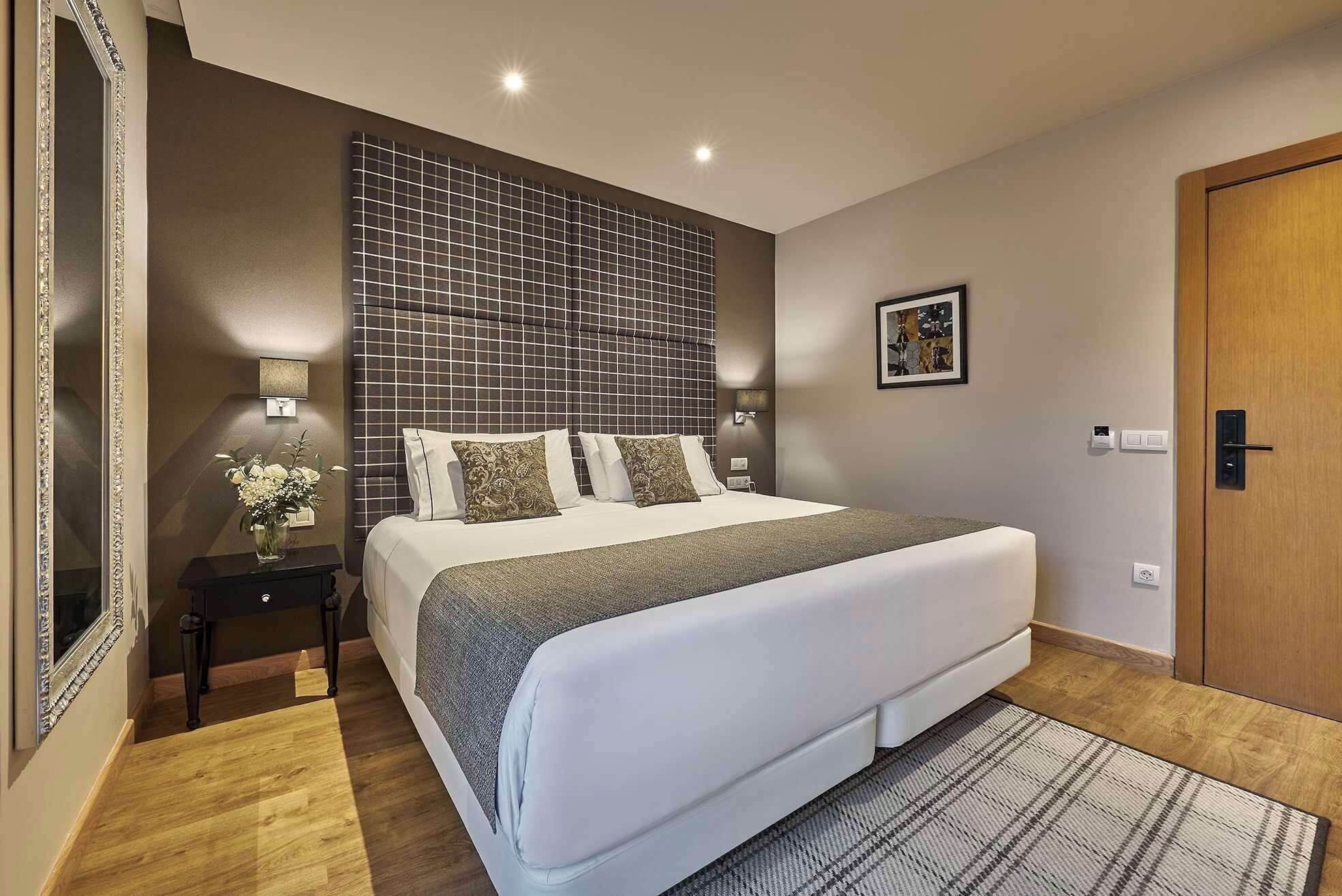 A modern hotel room with a large bed, a nightstand with a lamp, a wall-mounted TV, and a door.