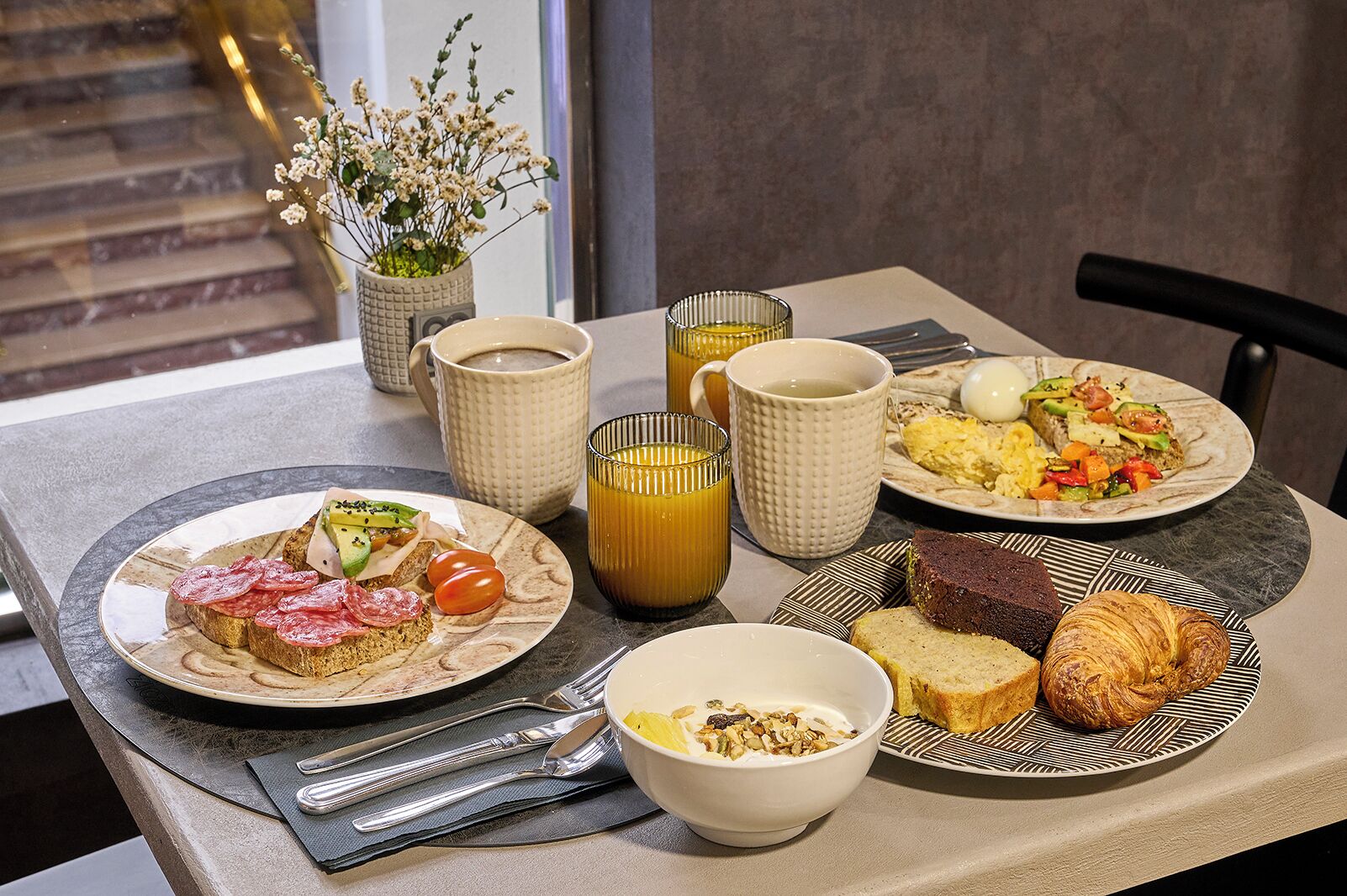 A breakfast table with plates of food, cups, and a vase with flowers.