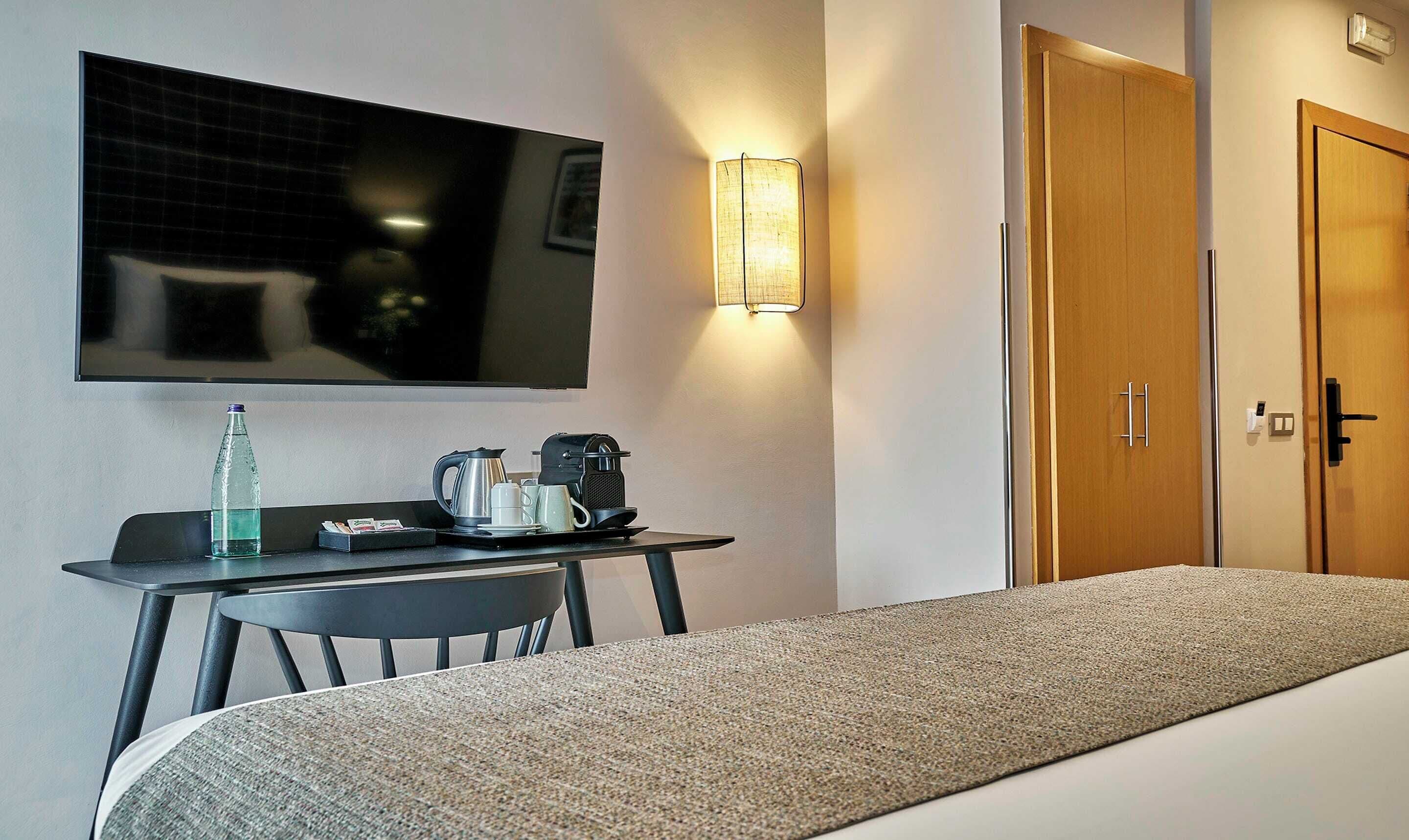 Una habitación de hotel con una cama, un escritorio con cafetera, una botella de agua y un televisor montado en la pared.