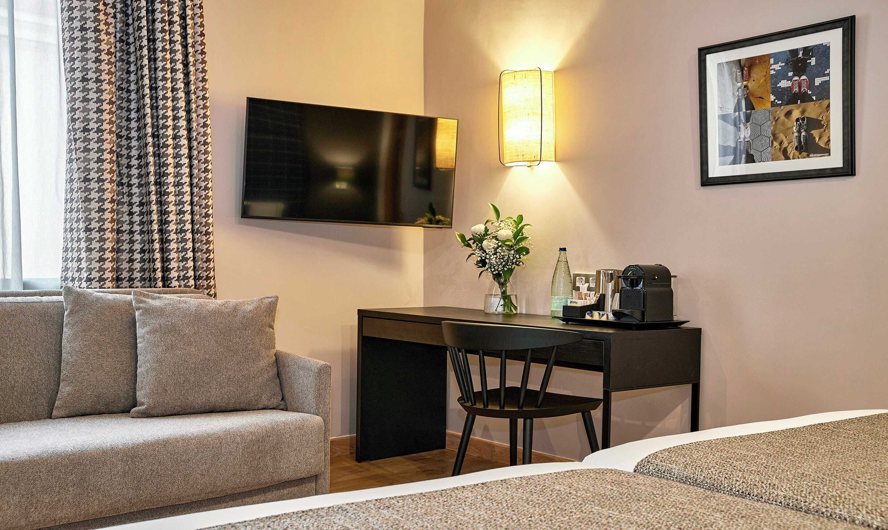 Habitación de hotel con sofá, escritorio y silla, televisión, lámpara de pared, una obra de arte enmarcada y una mesita con una botella y una cafetera.