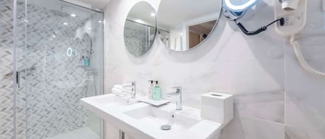 A modern bathroom with a glass shower enclosure, two sinks, and a hairdryer mounted on the wall.