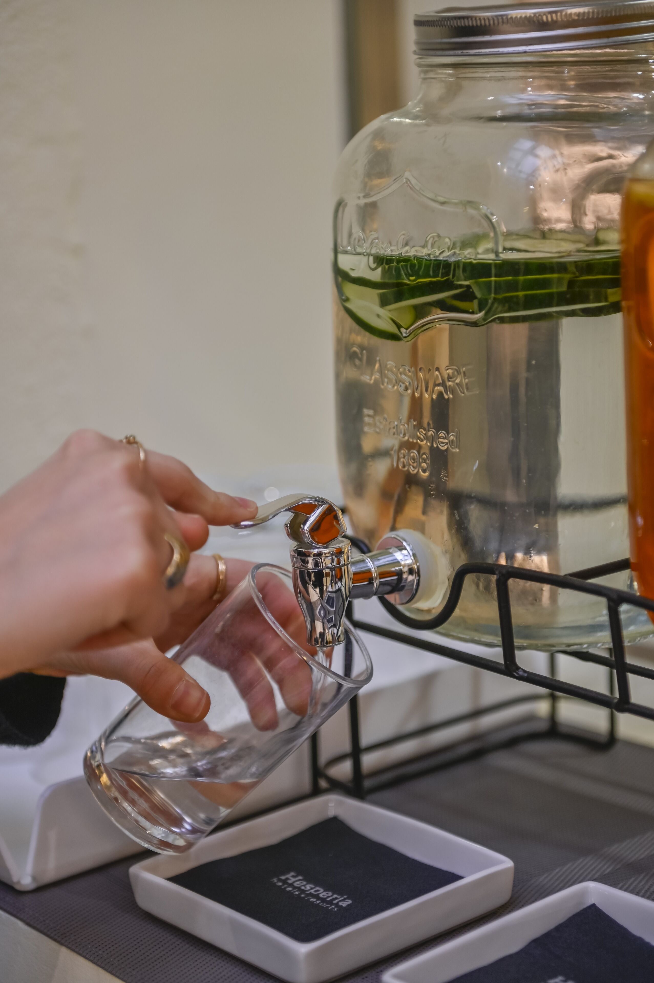 A hand filling a glass from a tap next to a large glass jar labeled 'GLASSWARE Established 1893'.