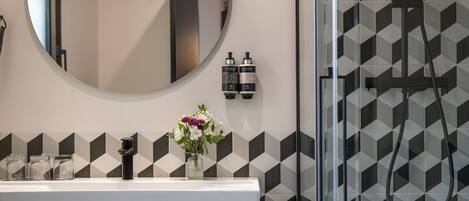 A bathroom with a modern design, featuring a round mirror, a sink with a vase of flowers, and a shower area with a geometric tile pattern.