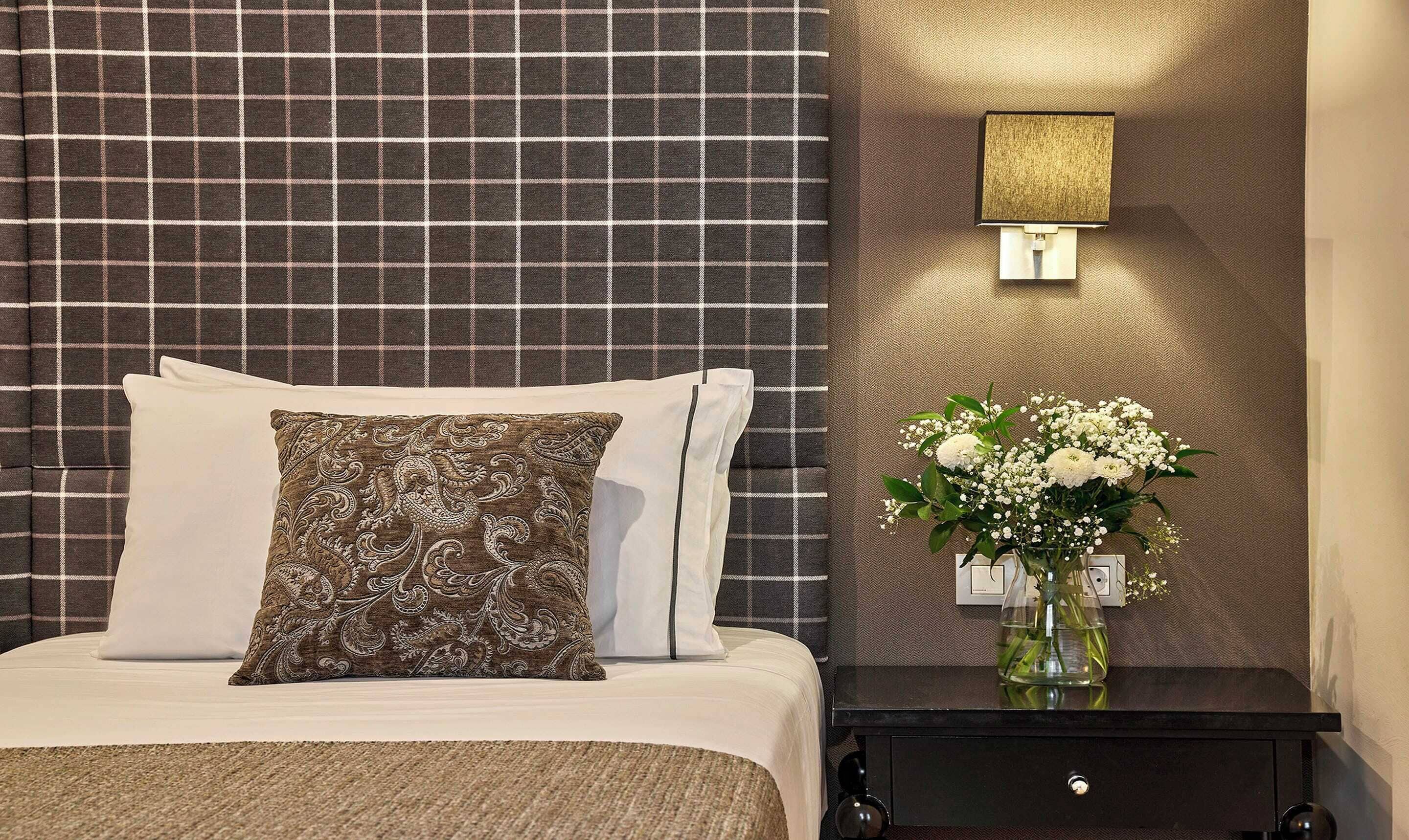 A neatly made bed with a patterned pillow, a bedside table with a vase of flowers, and a wall-mounted lamp.