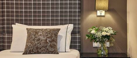 A neatly made bed with a patterned pillow, a bedside table with a vase of flowers, and a wall-mounted lamp.