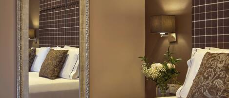 A hotel room with a large mirror, a bed with patterned pillows, a vase with flowers, and a wall-mounted light fixture.