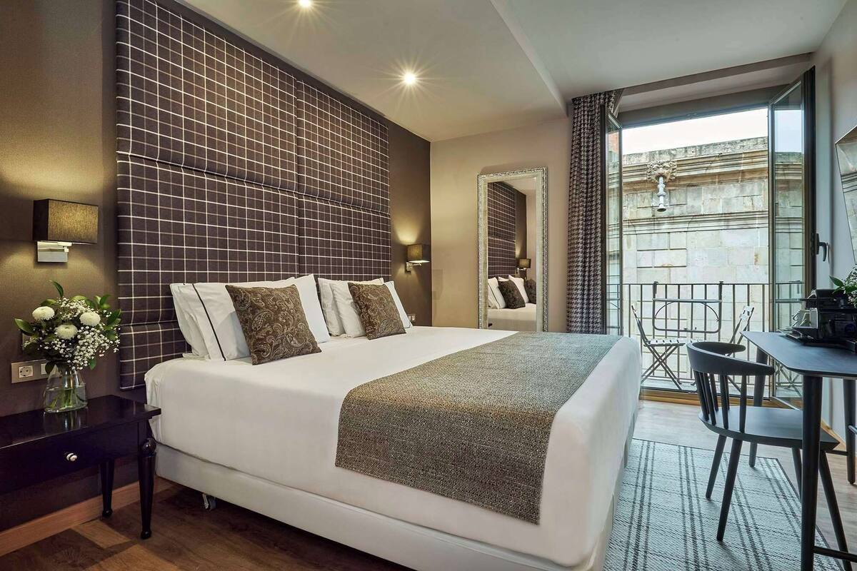 A hotel room with a large bed, a small table with chairs, a mirror, and a view of the city through a large window.