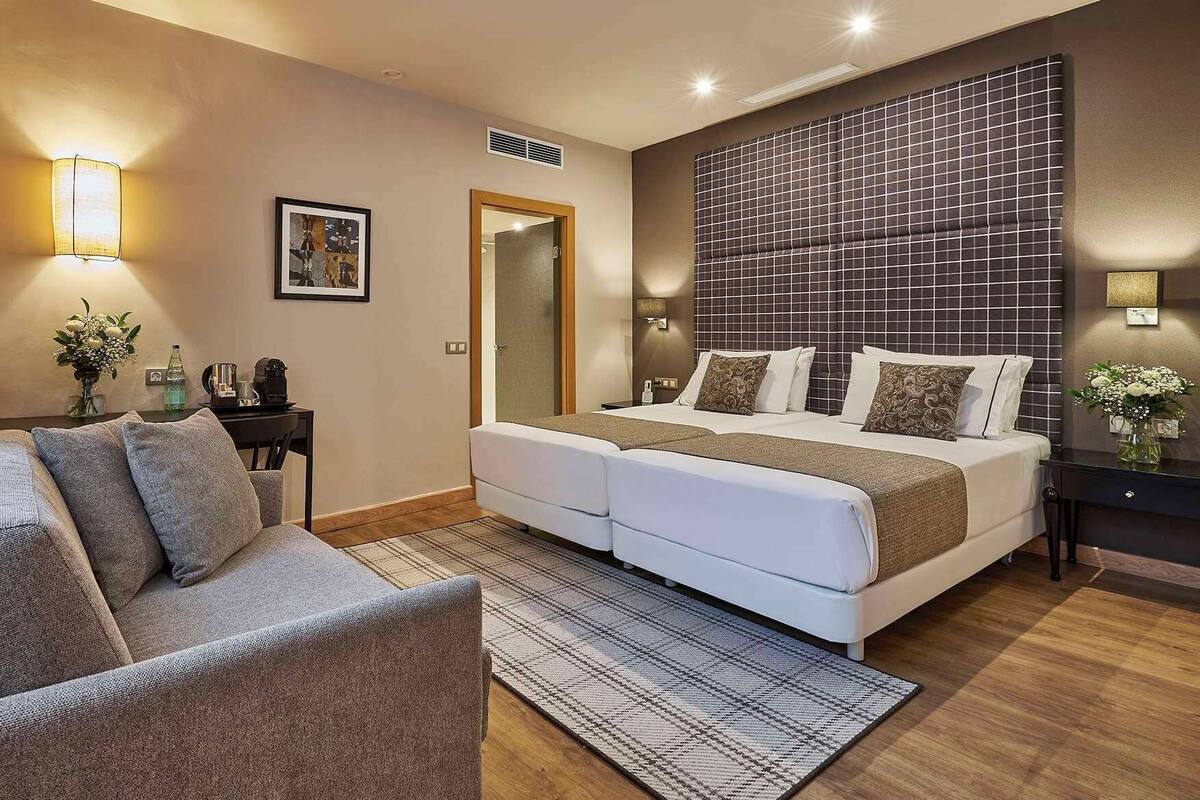 A modern hotel room with a large bed, a sofa, a desk with a lamp, and a mirror.