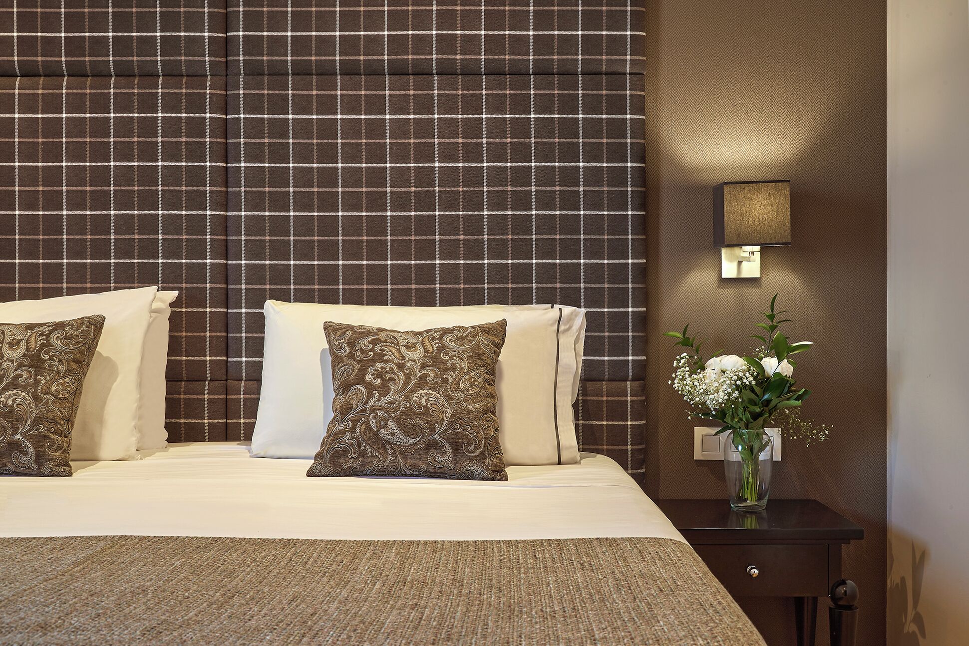 Una cama bien hecha con dos almohadas, una mesita de noche con un florero y una pared con un diseño texturizado.