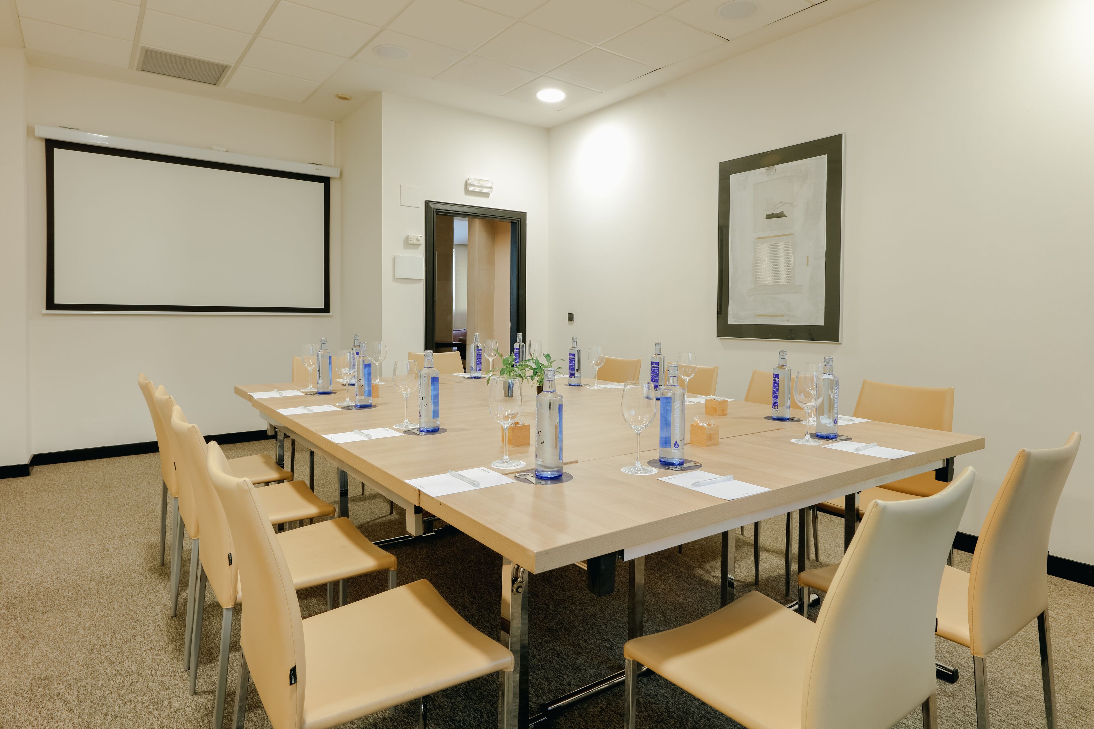 Ein Konferenzraum mit einem langen Tisch, Stühlen, einer Projektionsleinwand und einem gerahmten Aushang an der Wand.