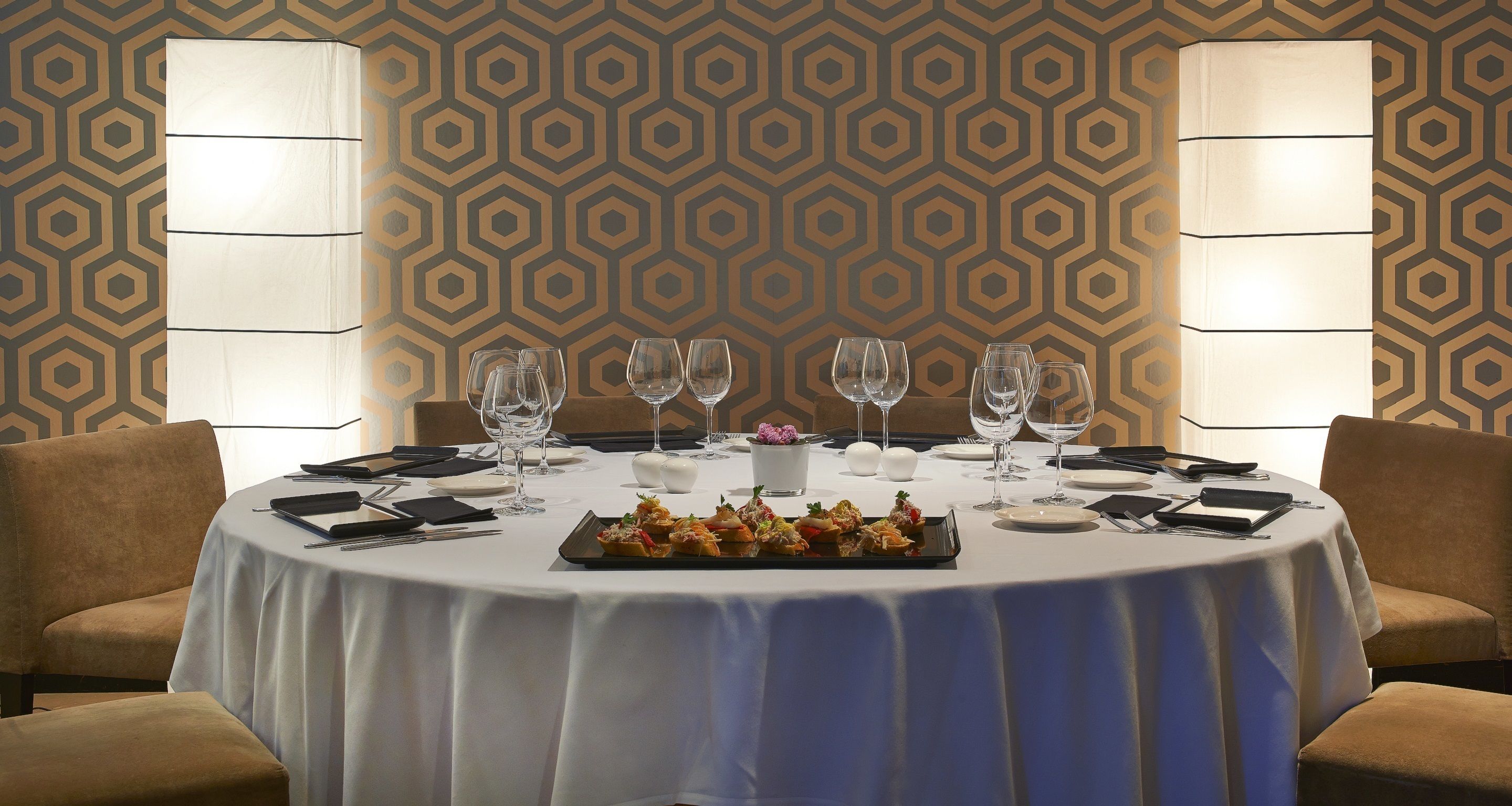 A round dining table with a white tablecloth, surrounded by four brown chairs. There are six wine glasses and a small floral centerpiece on the table.