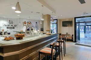 Café-bar com balcão exibindo comida e bebidas, banquetas e parede com obras de arte.