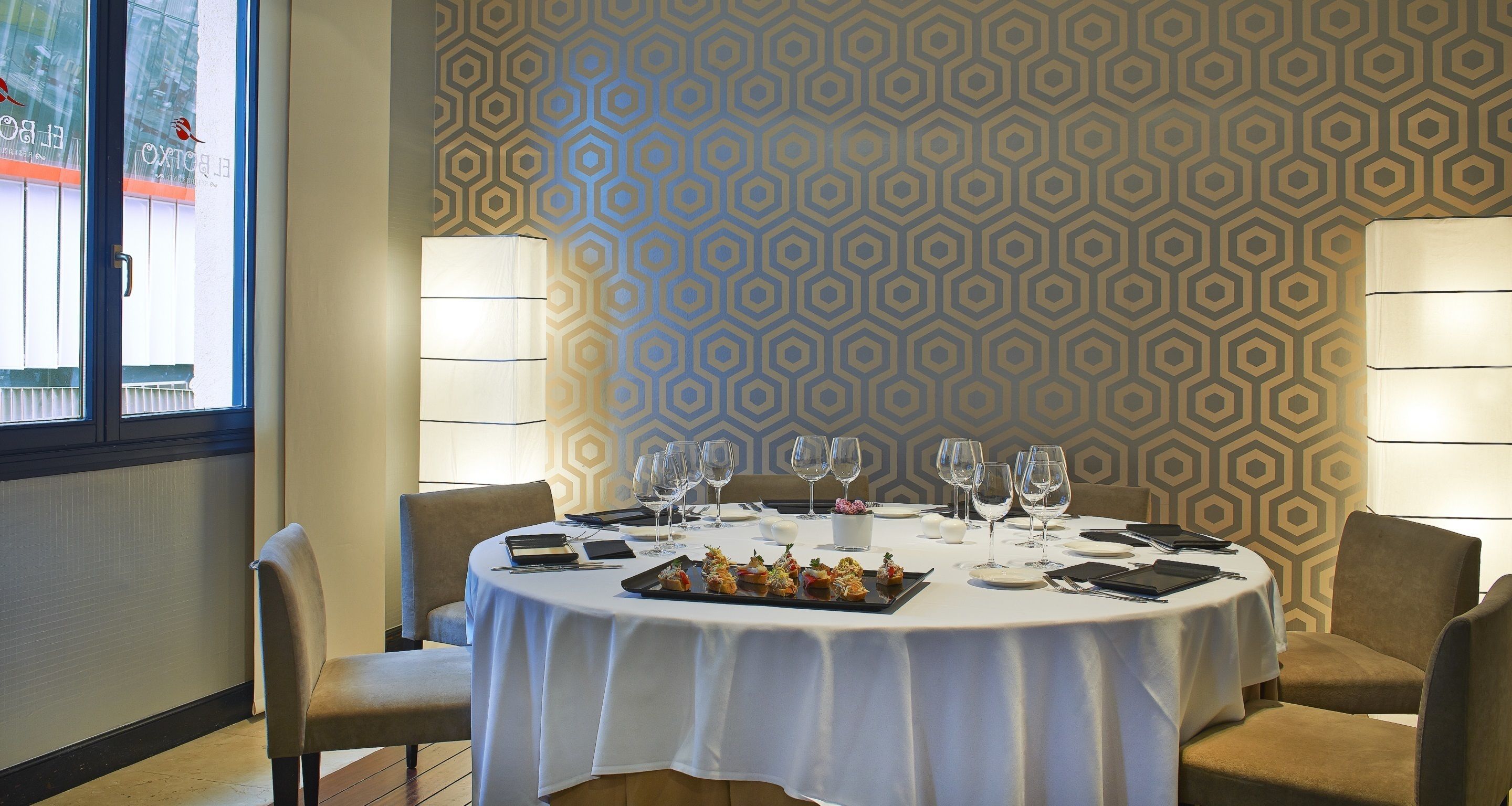 Una mesa redonda para cuatro personas con mantel blanco, rodeada por cuatro sillas. Hay platos, cubiertos y vasos sobre la mesa. La habitación tiene un papel tapiz con diseño y dos lámparas fijadas a la pared.