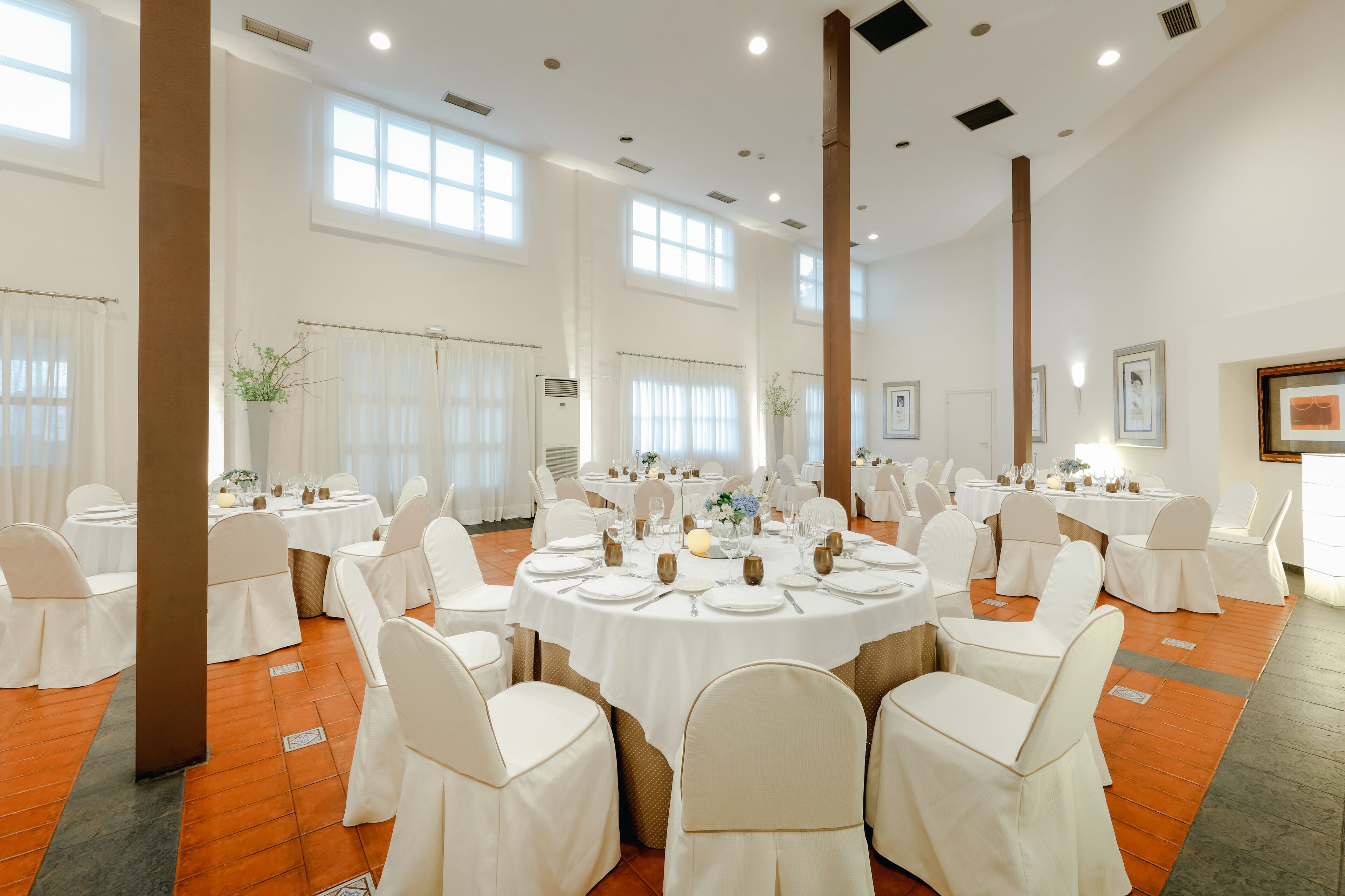 A banquet hall with round tables set for dining, white chairs, and large windows allowing natural light.