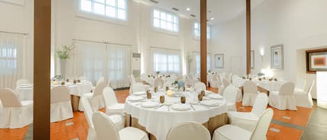 A banquet hall with round tables set for dining, white chairs, and large windows allowing natural light.