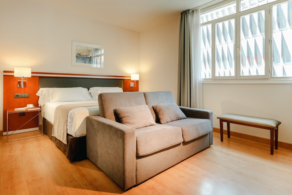 Una habitación de hotel con una cama, dos mesitas de noche, un sofá, un banco y una ventana con persianas.