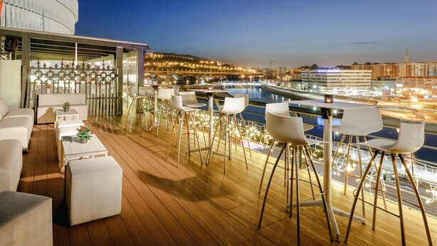 A deck with white furniture, a pool, and city lights in the background. - Abba Euskalduna Hotel (Bilbao)