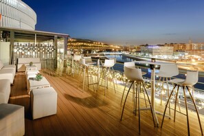 Um terraço com móveis brancos, piscina e luzes da cidade ao fundo.